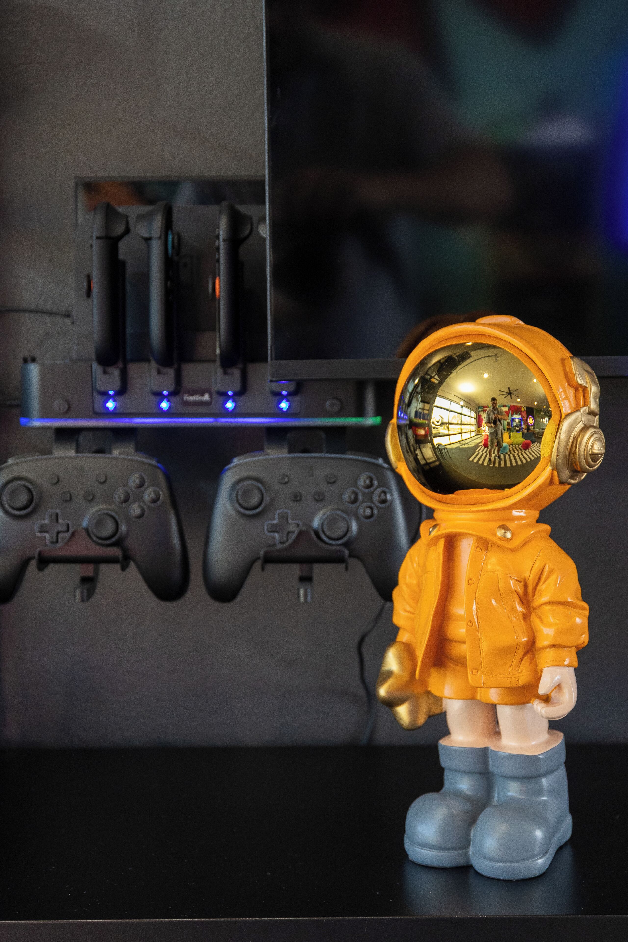 A figurine of an astronaut with a helmet visor reflecting a room with gaming controllers and a blue-lit shelf.