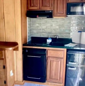 Eine kompakte Küche mit Holzausstattung, einem schwarzen Geschirrspüler, einem Edelstahlkühlschrank und einer Mikrowelle.