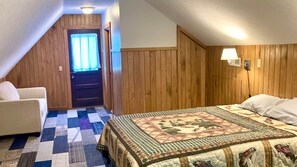 Ein Schlafzimmer mit holzverkleideter Schräge, einer gesteppten Bettdecke, einem Stuhl und einer Tür mit Fenster.