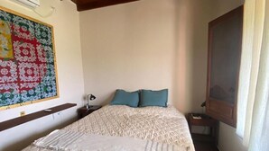 Un dormitorio con una cama, almohadas, una mesita de noche, una lámpara, un adorno en la pared y una ventana con cortinas.
