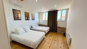 A bedroom with two beds, wooden flooring, an air conditioning unit, and a window with curtains. - Apartment Camille Pujol Toulouse (Toulouse)