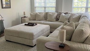 Ein Wohnzimmer mit Ecksofa, Hocker, einem kleinen Tisch und Deckenventilator.