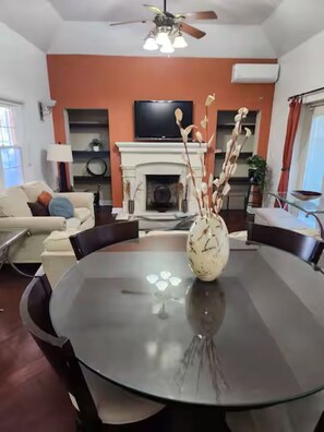 A modern living room with a round glass table, a fireplace, a ceiling fan, and a TV mounted on the wall. - Two-bedroom house that sleeps 6 in St. George's. Your home away from home! (St.George's)