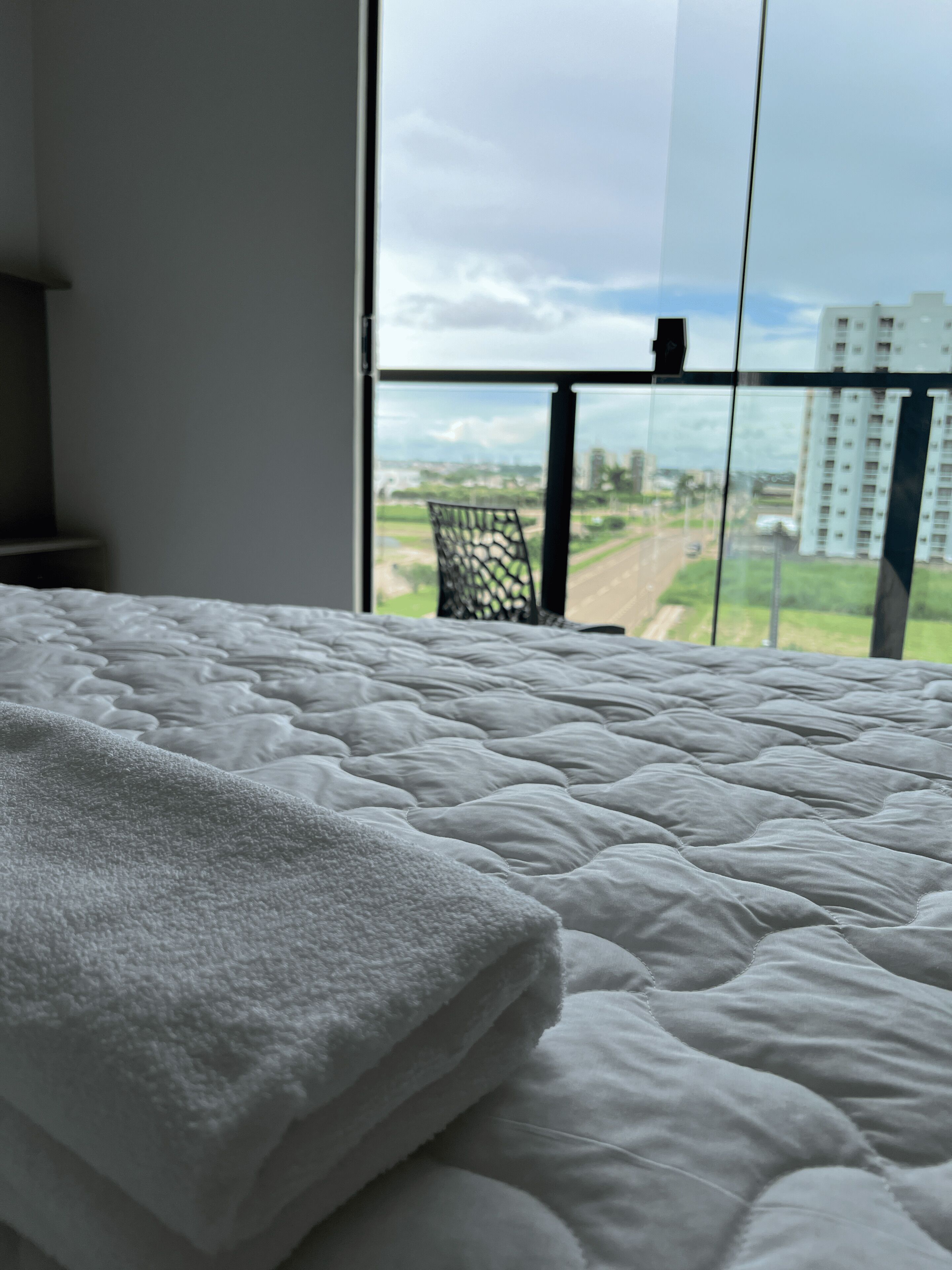 A bed with a white quilt and a view of a cityscape through a large window.