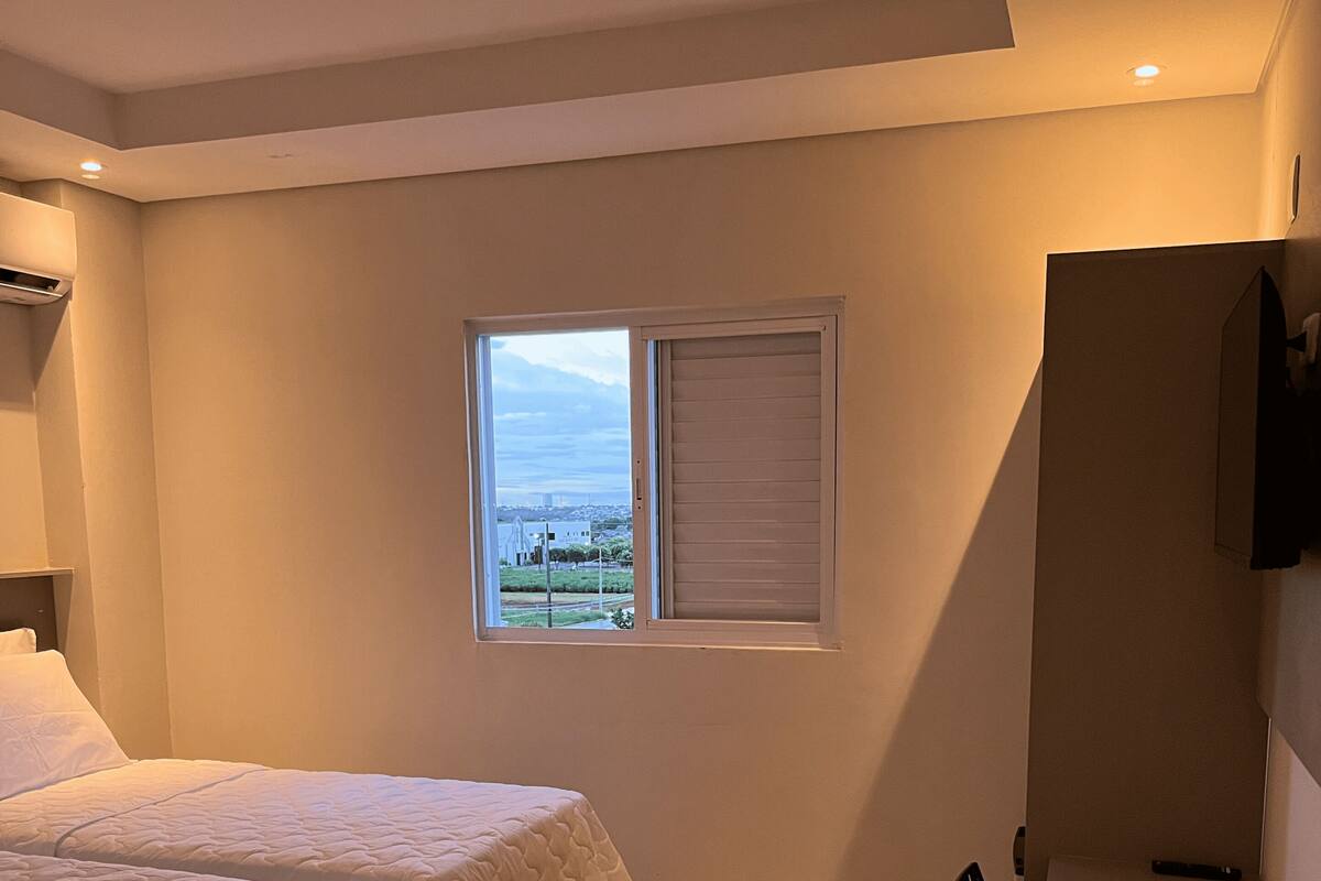 A bedroom with two beds, a window with blinds, a chair, and a television on the wall.
