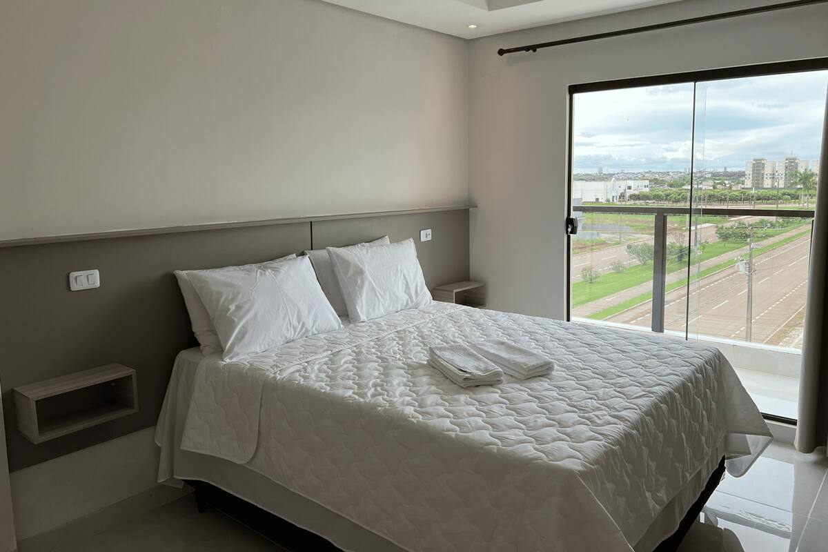 A bedroom with a large bed, a nightstand, a window with a view of a cityscape, and a balcony.