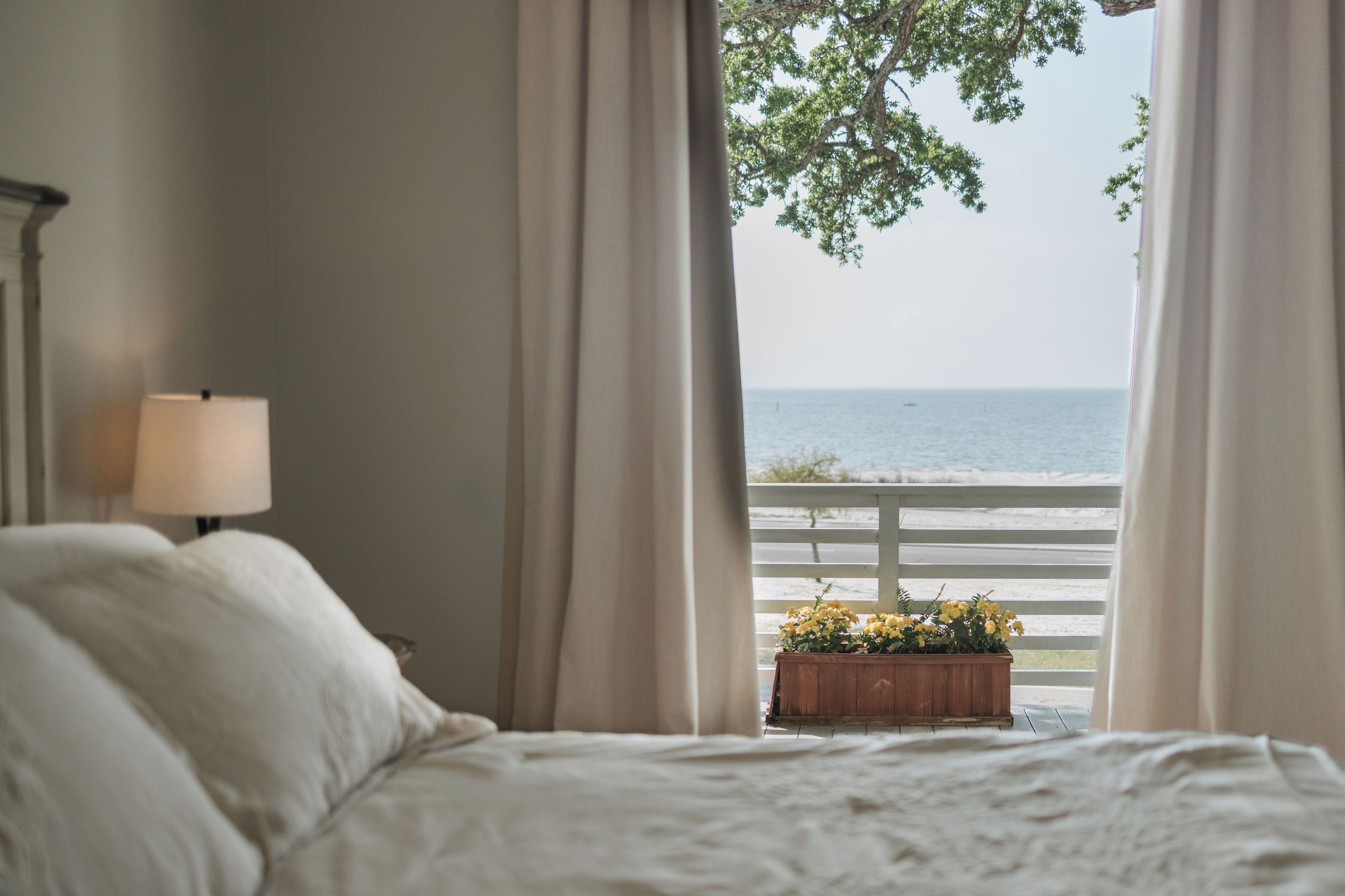 Une chambre avec un lit, une table de chevet avec une lampe, une vue sur la mer à travers les rideaux et un jardinière en bois avec des fleurs.