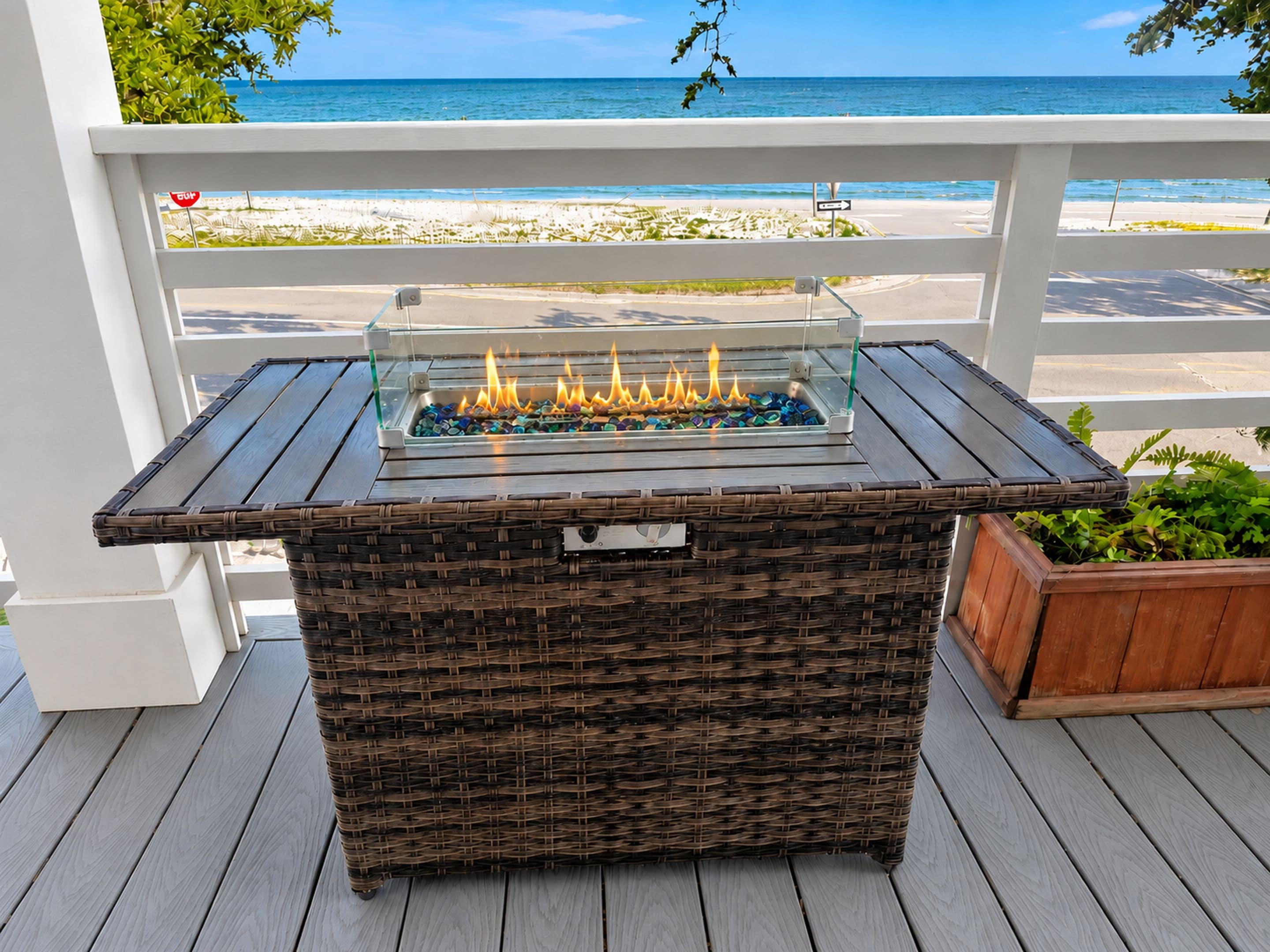 A wicker outdoor bar with a glass-top table and a built-in fire feature, overlooking a beach.