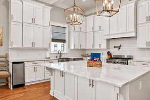 A modern kitchen with white cabinetry, a large island, a gas stove, and a pendant light fixture. - Gulf View Beach House / Sleeps 18 (Gulfport)
