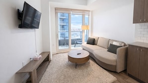 Ein modernes Wohnzimmer mit einer Couch, einem Couchtisch, einem an der Wand befestigten Fernseher und Blick ins Freie durch große Fenster.