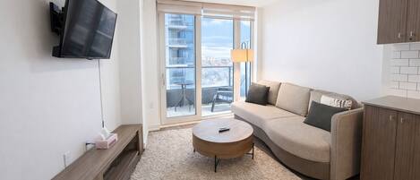 A modern living room with a sofa, a coffee table, a TV mounted on the wall, and a view of the outside through large windows.