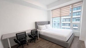 Ein Schlafzimmer mit einem Bett, zwei Bürostühlen, einem Schreibtisch und einem großen Fenster mit Jalousien.