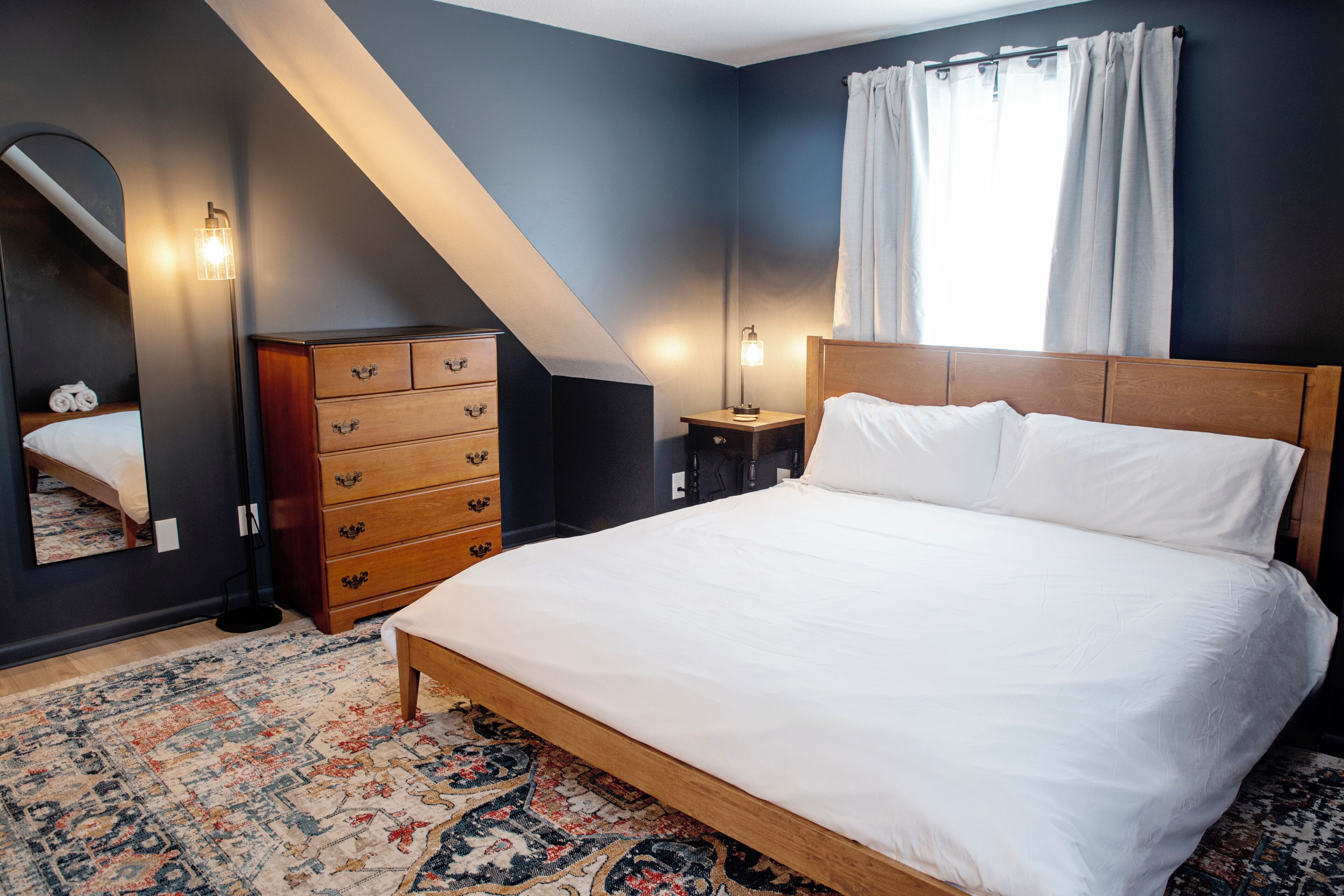 Ett sovrum med en stor säng, en byrå, ett nattduksbord, en spegel och ett fönster med gardiner.