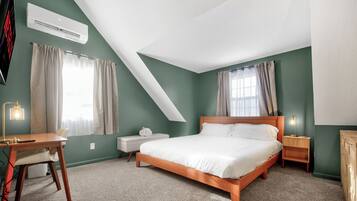A bedroom with a bed, a desk, a chair, a nightstand, and an air conditioning unit.