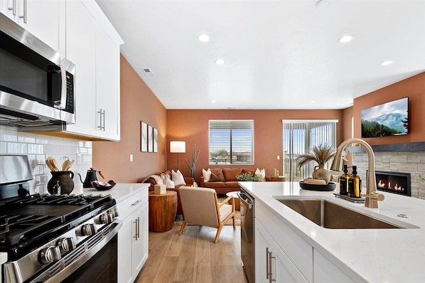 A modern kitchen with a stove, microwave, and sink; a living area with a sofa and fireplace; and a large window with blinds.