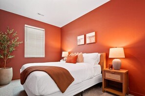 A bedroom with a bed, bedside lamps, a potted plant, and two framed pictures on the wall.