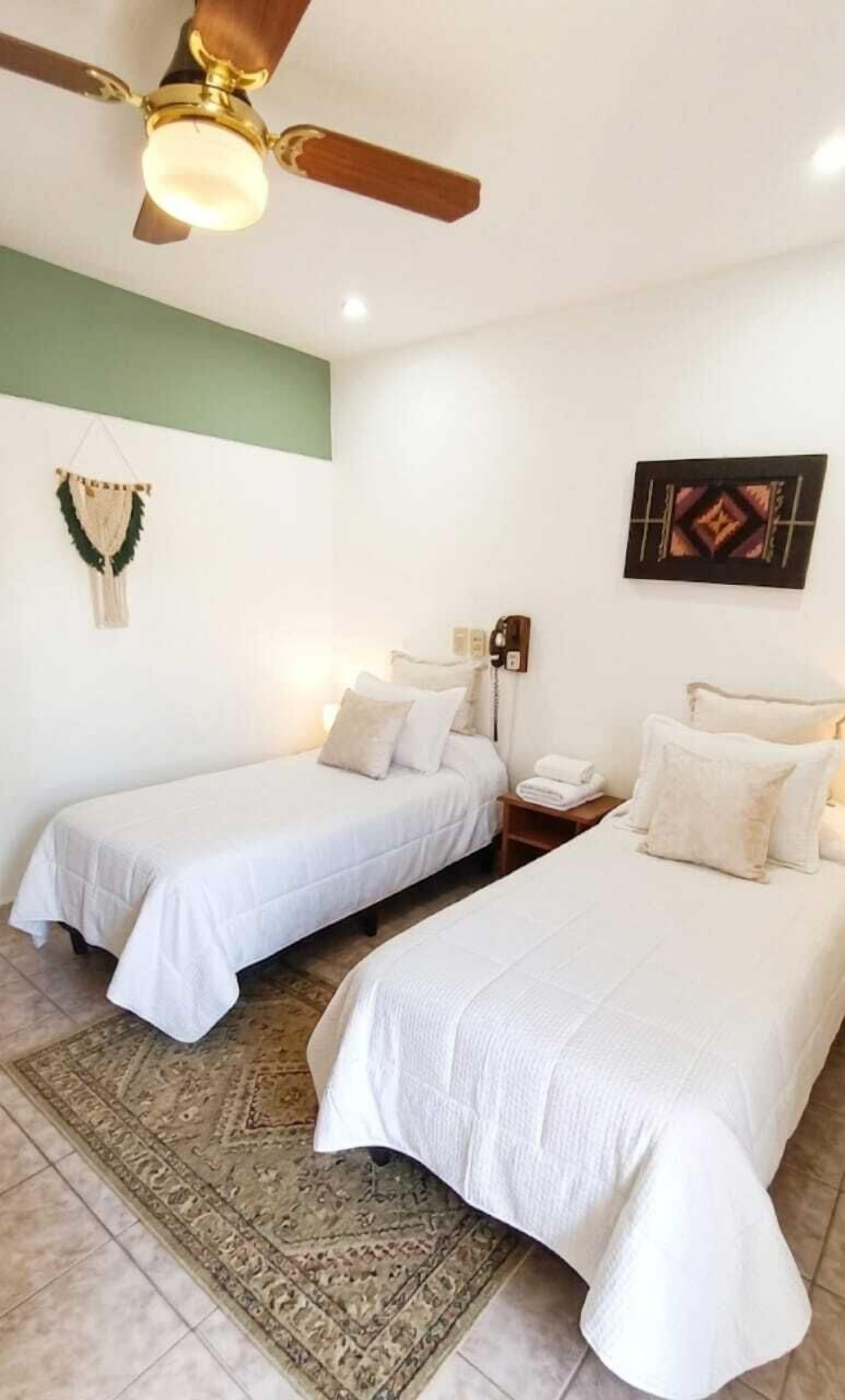 A hotel room with two single beds, a ceiling fan, a wall-mounted phone, and a decorative wall hanging.