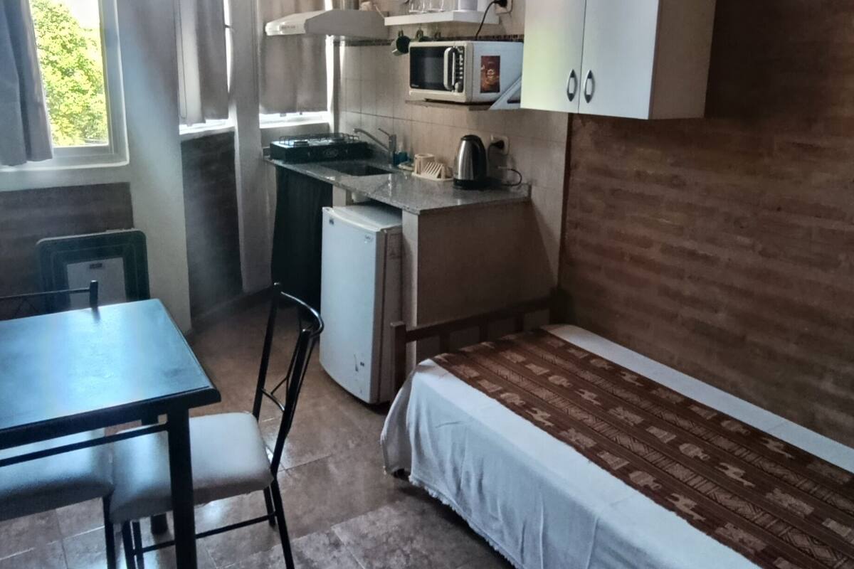 Een compacte keuken met een magnetron, spoelbak en een kleine koelkast. Er is een eettafel met stoelen en een bank.