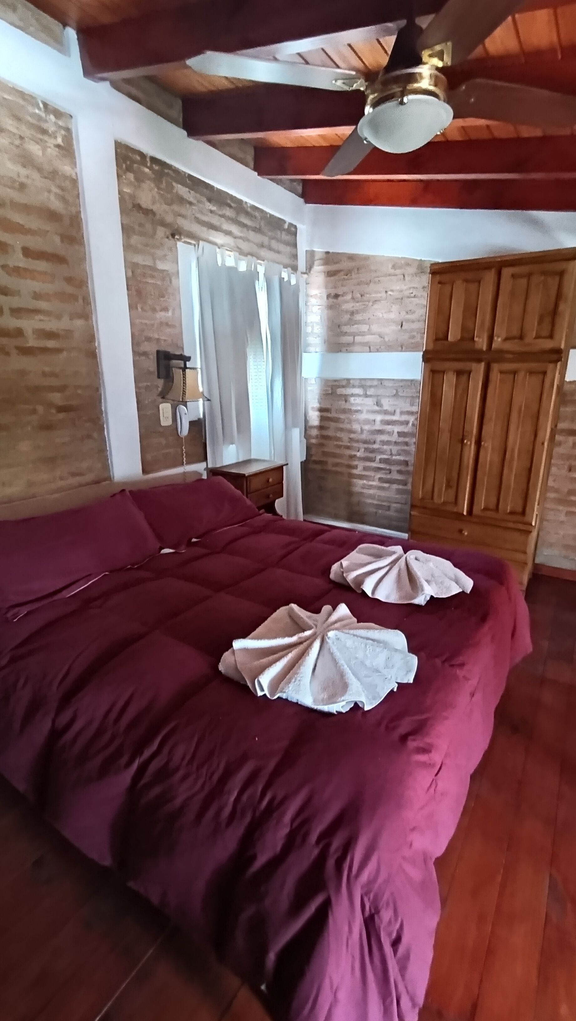 A room with a wooden ceiling, a bed with a red bedspread, a wooden wardrobe, and a ceiling fan.
