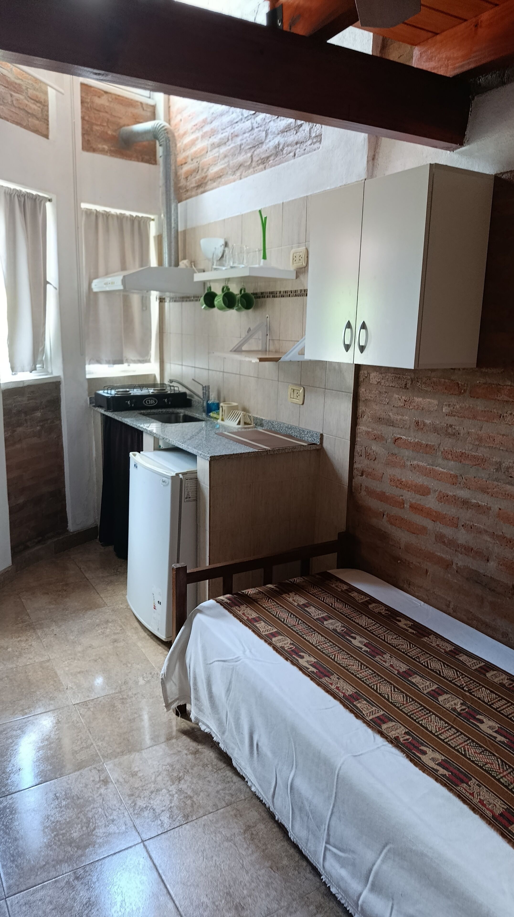 A compact kitchen with a built-in stove, a sink, and a small refrigerator. There is a window with curtains, and the room has a tiled floor.