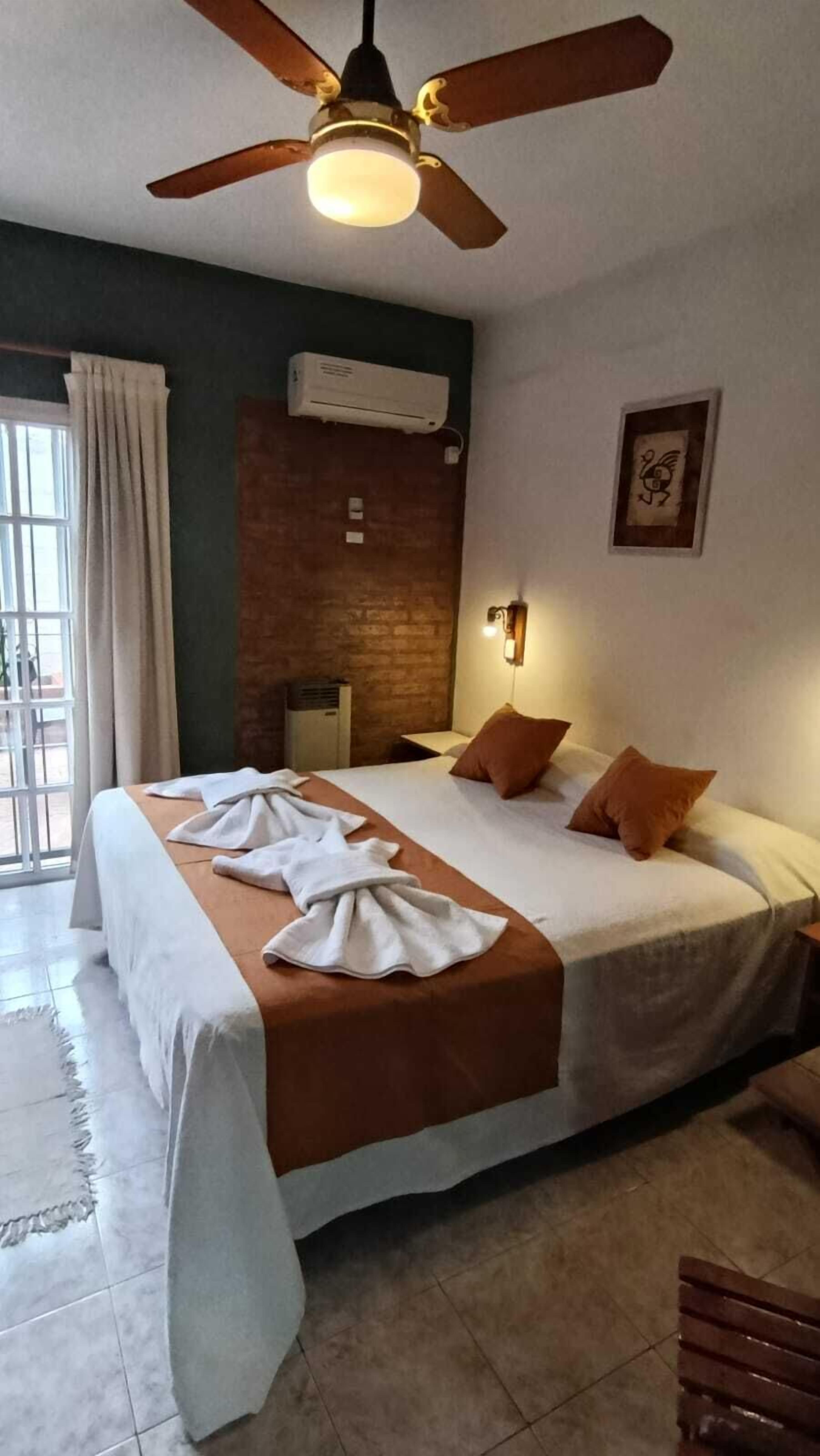 A hotel room with a bed, a ceiling fan, an air conditioning unit, a wall-mounted light, and a window with curtains.