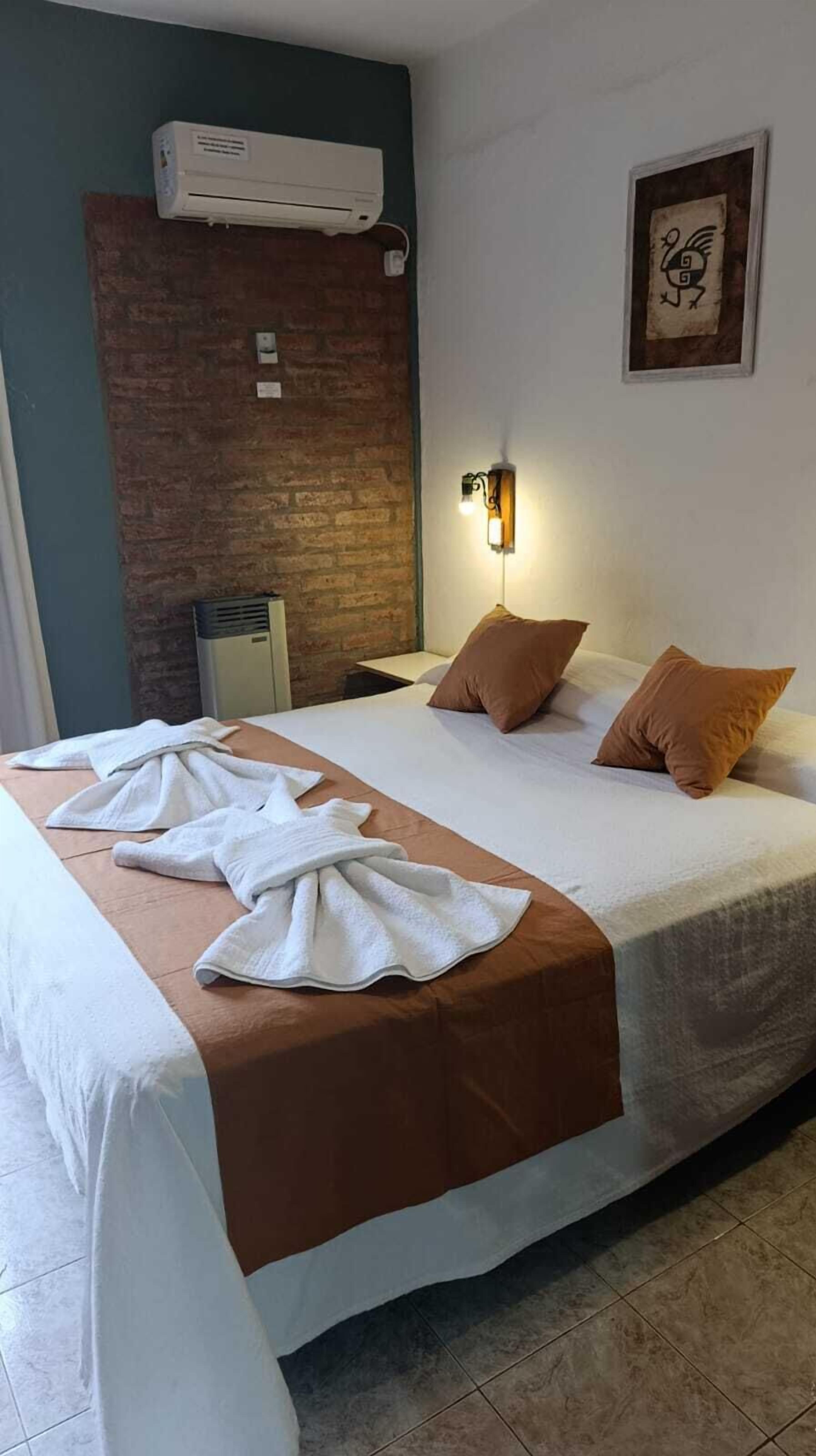 A hotel room with a bed, a brick wall, an air conditioning unit, a wall-mounted light, and a framed picture.