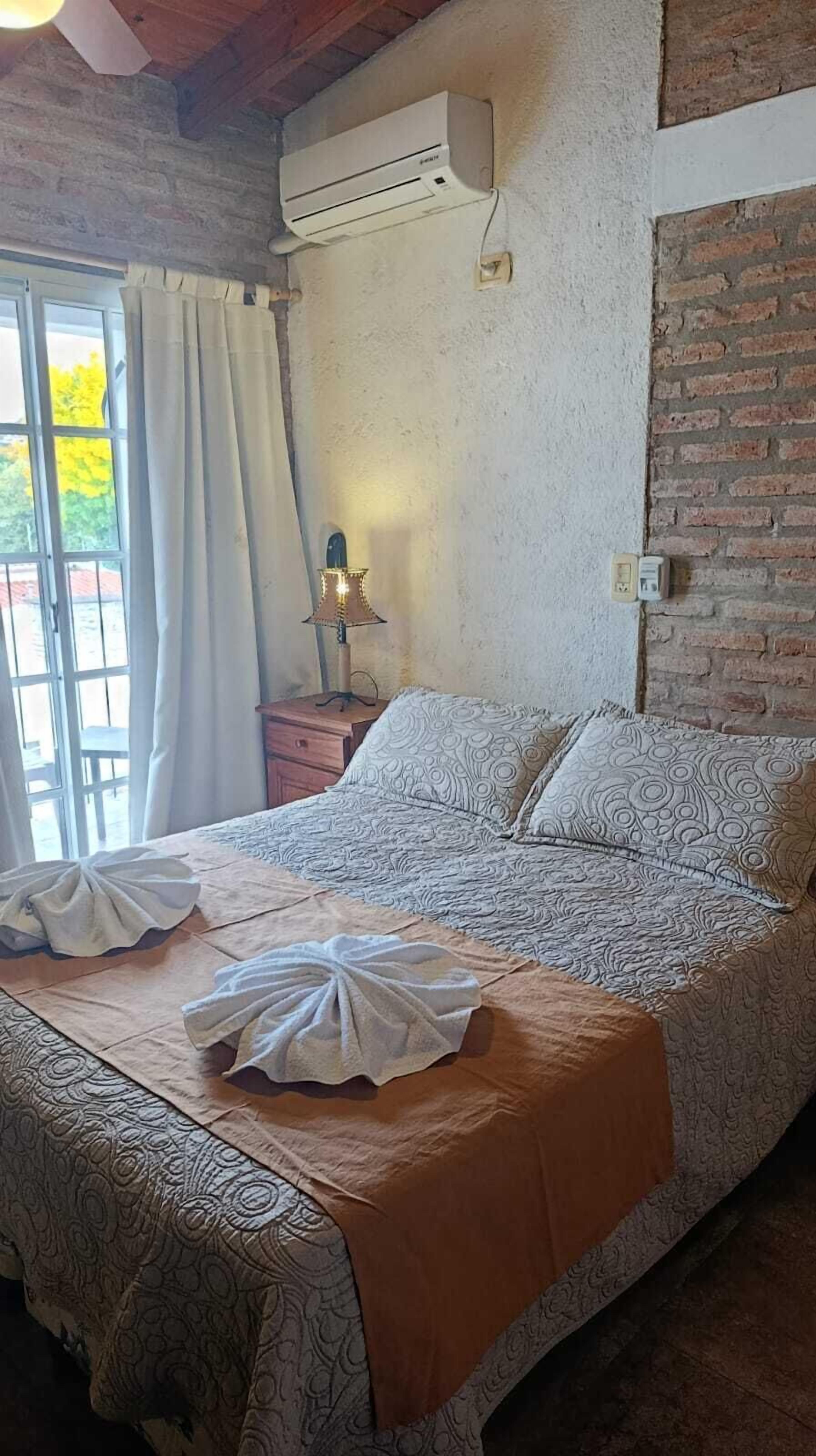 Een slaapkamer met een bed, nachtkastje, airconditioning, lamp en raam met gordijnen.