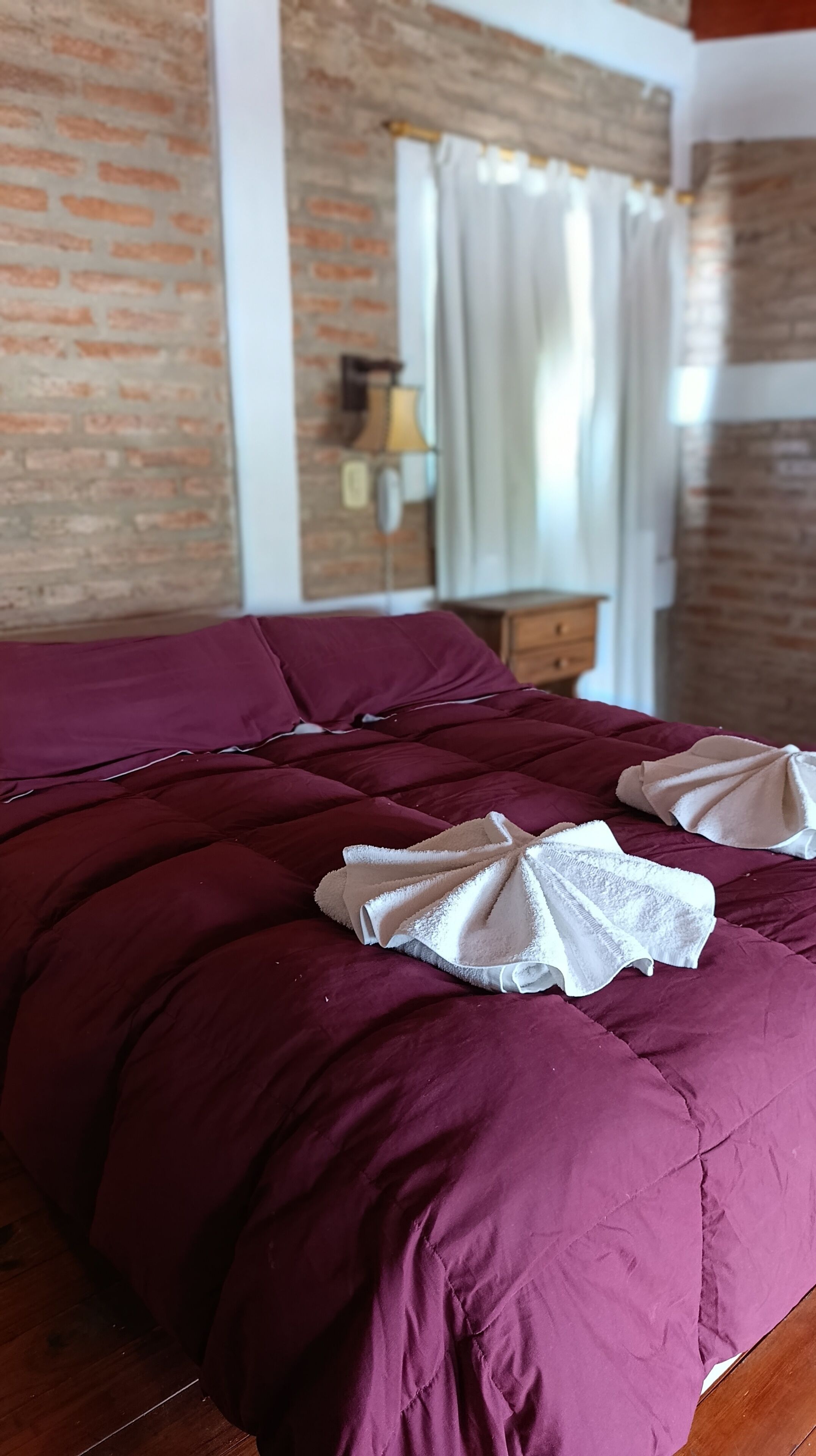 A bed with a purple bedspread and two white towels, a brick wall, a window with curtains, and a nightstand with a lamp.