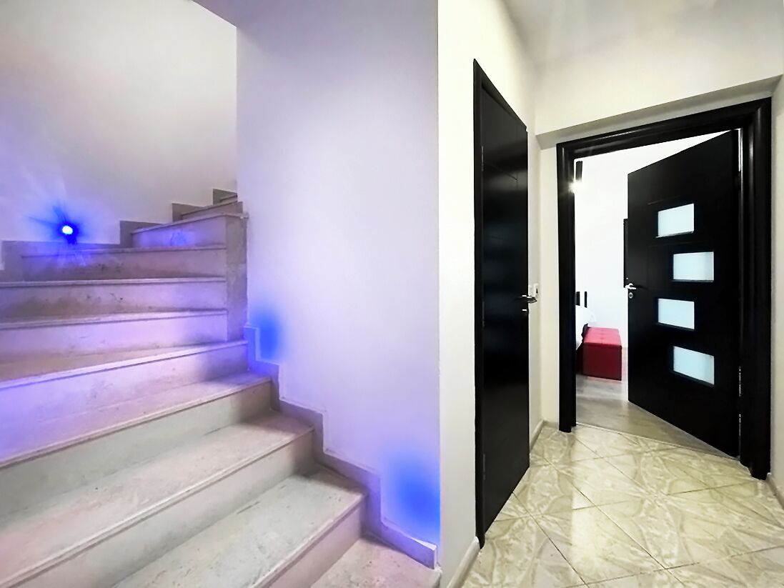 Een trap met blauw licht bovenin en een zwarte deur met een rode bank in de hal.