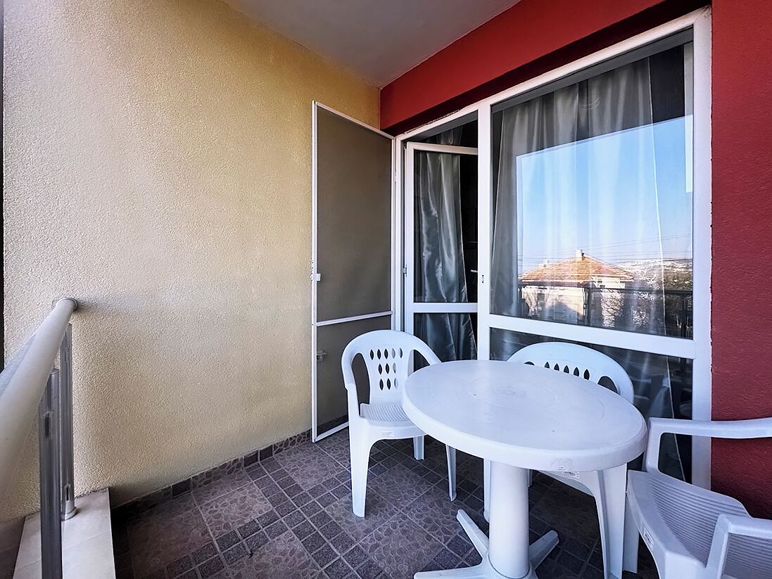 Un balcó amb taula i cadires blanques, una porta corredissa de vidre i vista a un edifici.