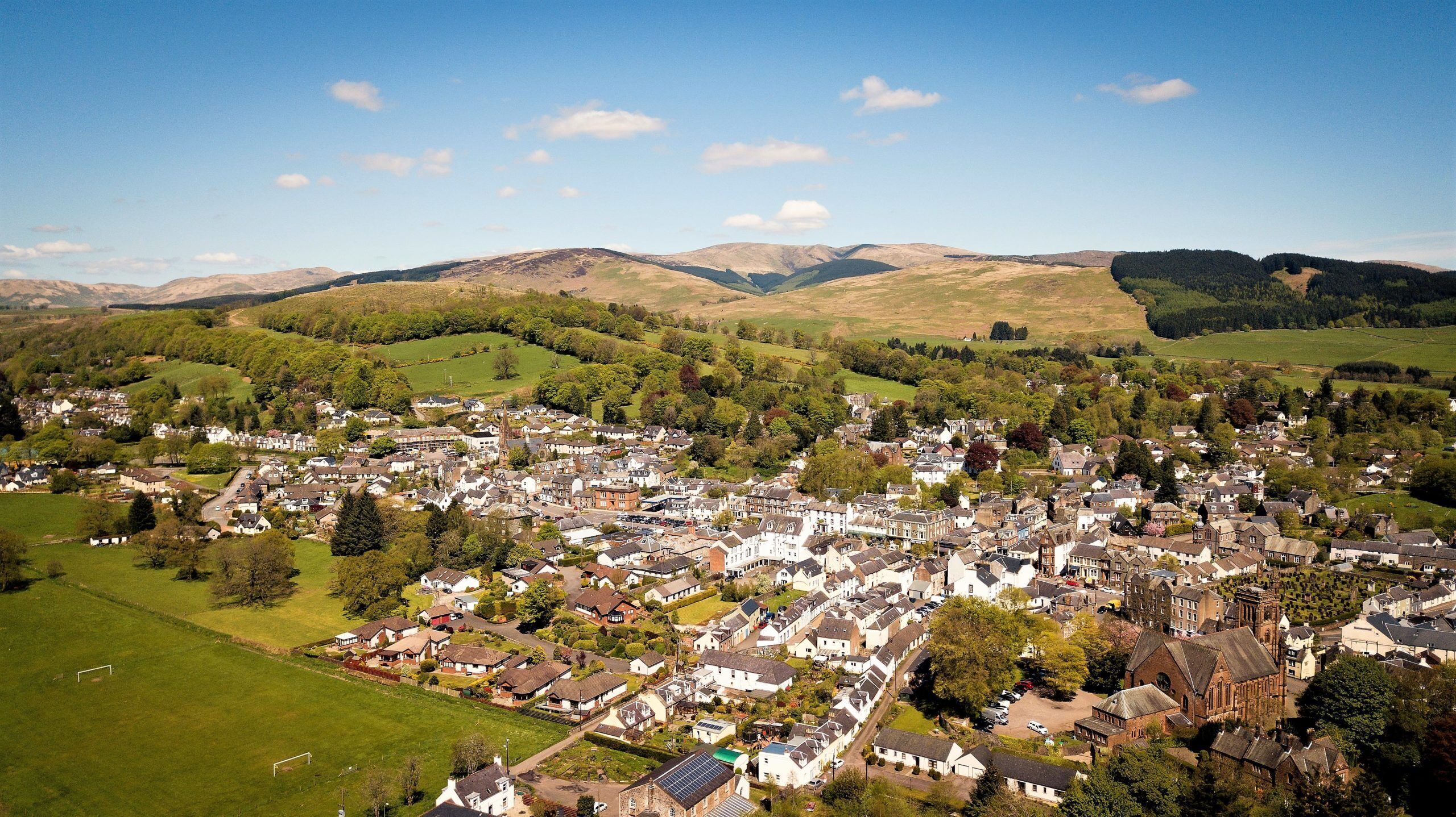 A town with a mix of residential and commercial buildings, surrounded by green hills and a clear sky.