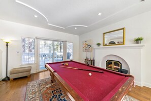 Une table de billard dans une pièce avec une cheminée, un banc et une vue sur l’extérieur grâce à de grandes fenêtres.