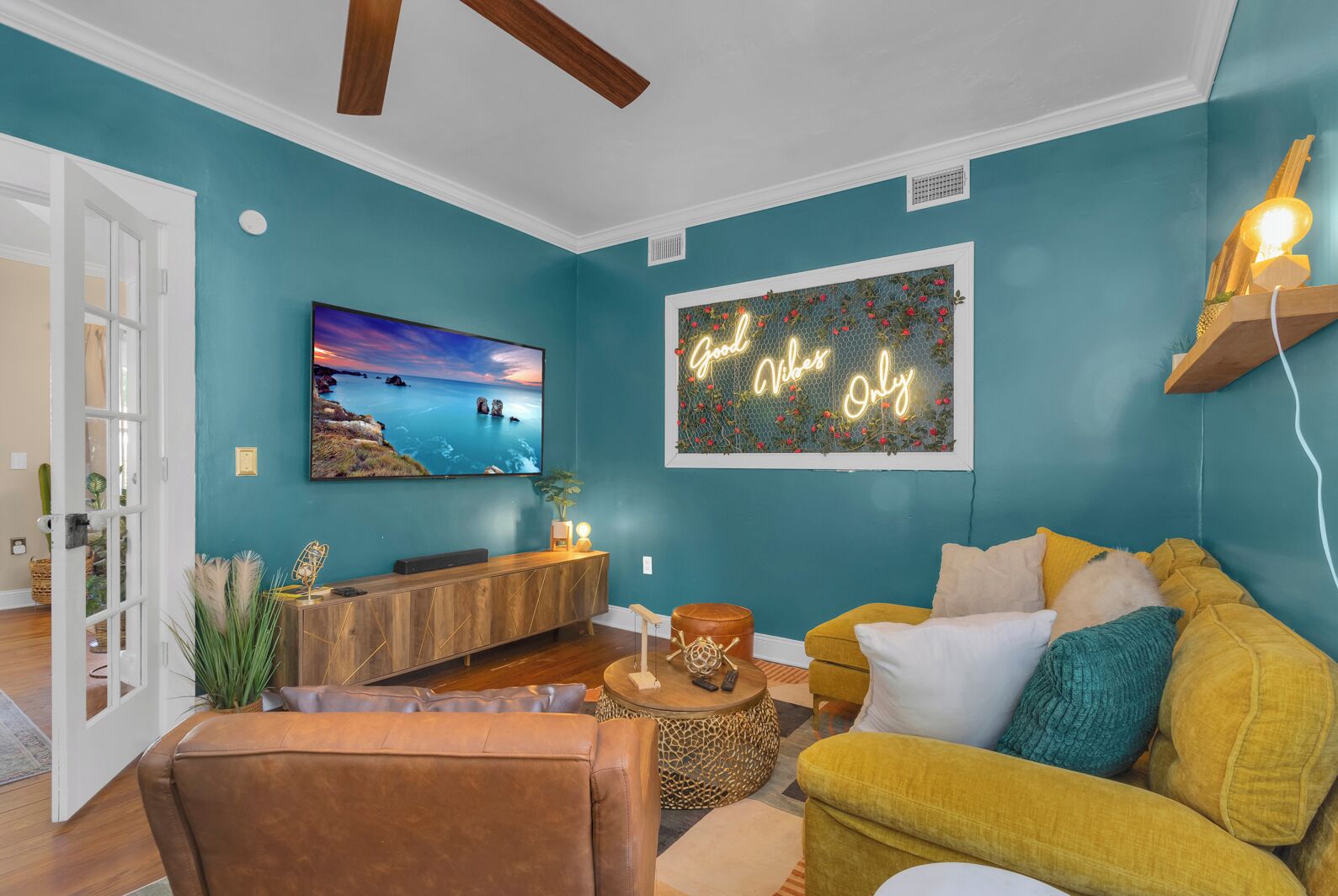A living room with teal walls, a mounted TV, a neon sign, and a yellow sofa.