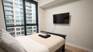 Une chambre à coucher avec un lit, un téléviseur fixé au mur et une fenêtre donnant sur un immeuble.