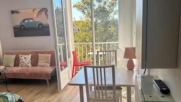 A room with a wooden floor, a white table with a chair, a pink sofa, a TV, and a balcony with a view of trees and a car. - City center studio (Vendôme)