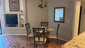 Un coin repas comprenant une table ronde, quatre chaises, un lustre, un téléviseur et une cheminée surmontée d’un tableau encadré.