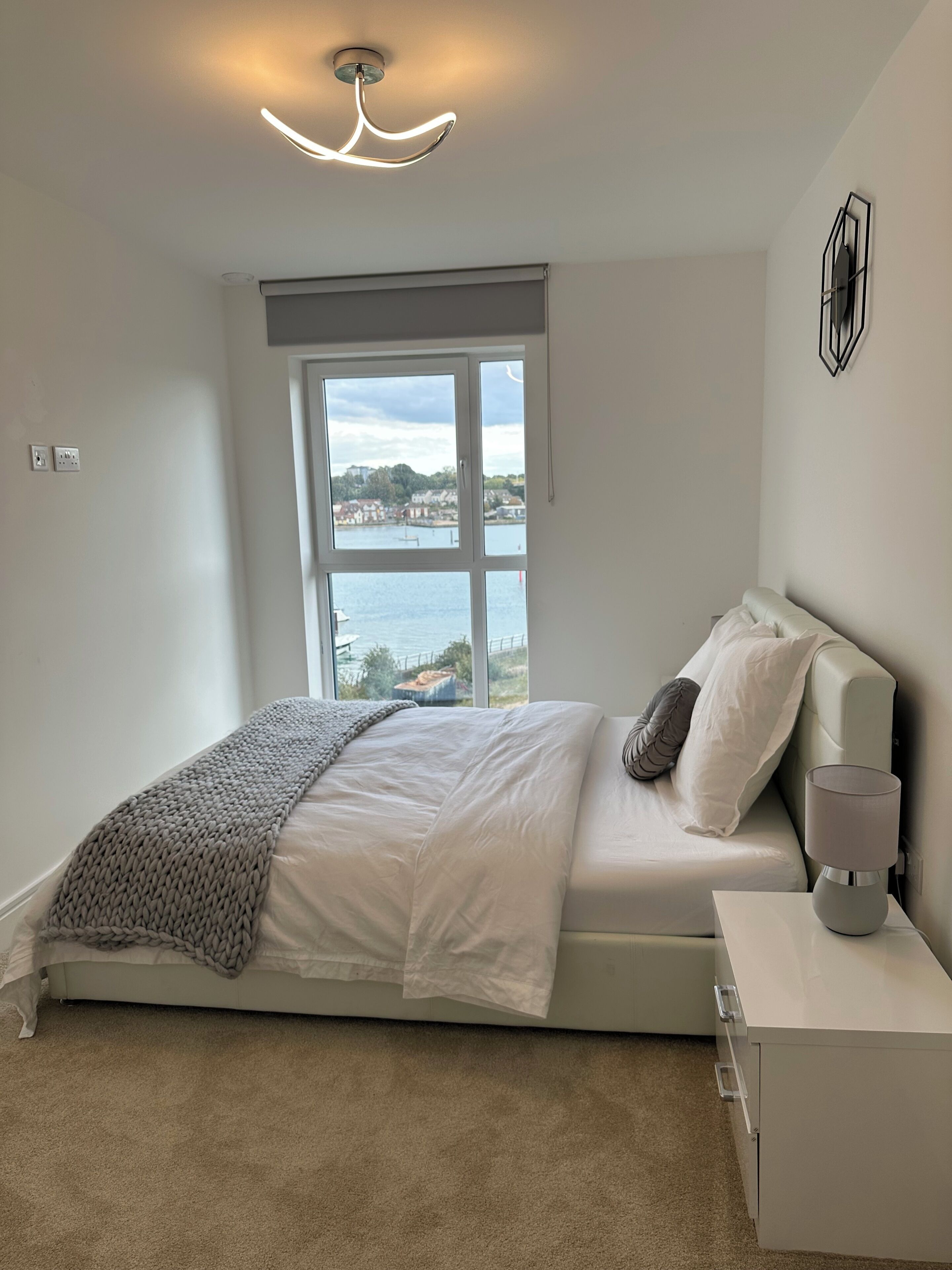 Een slaapkamer met een bed, nachtkastje, lamp, raam met uitzicht op water en gebouwen, en een plafondlamp.