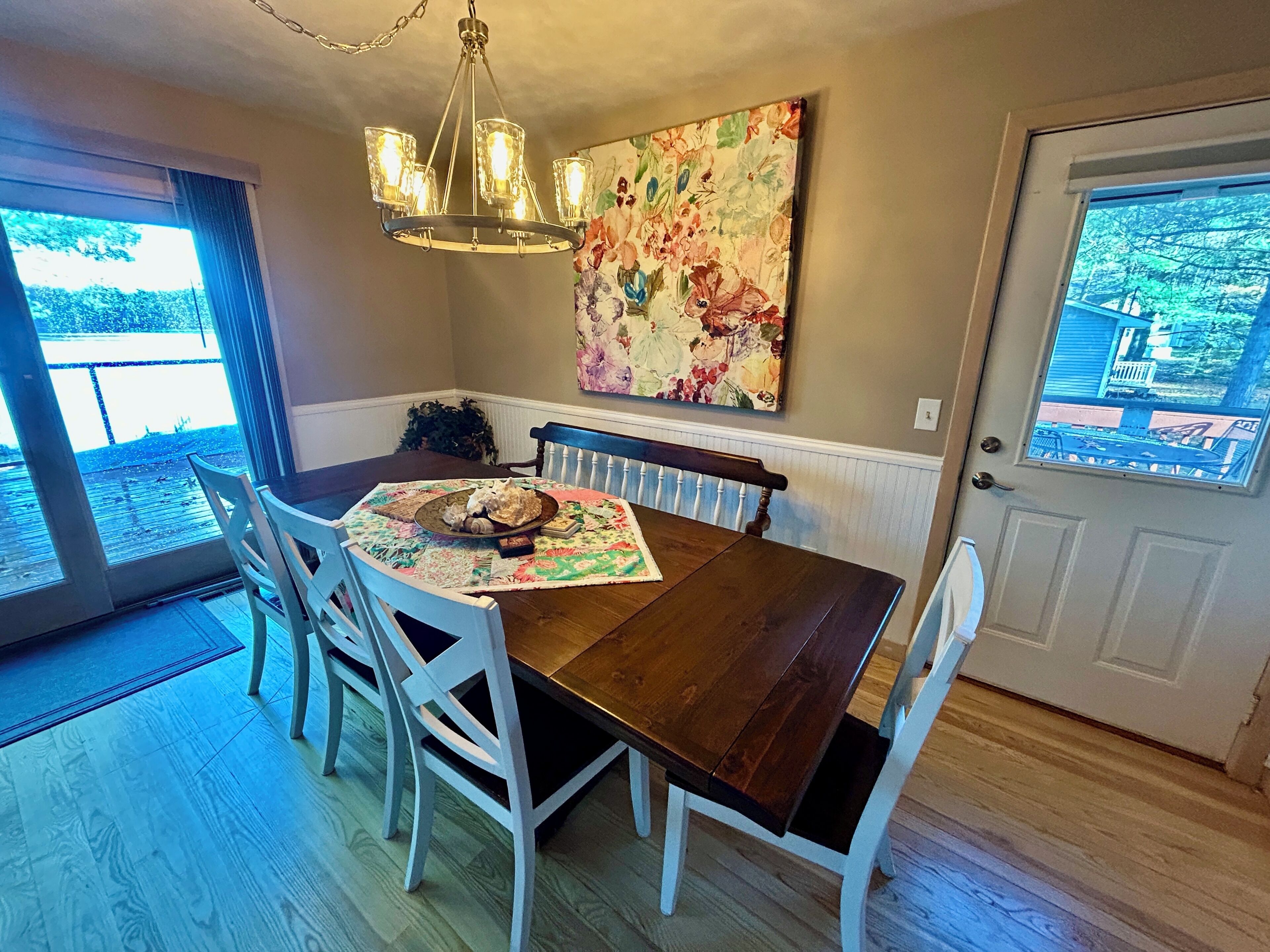 A dining area with a wooden table, white chairs, a chandelier, and a floral painting on the wall.