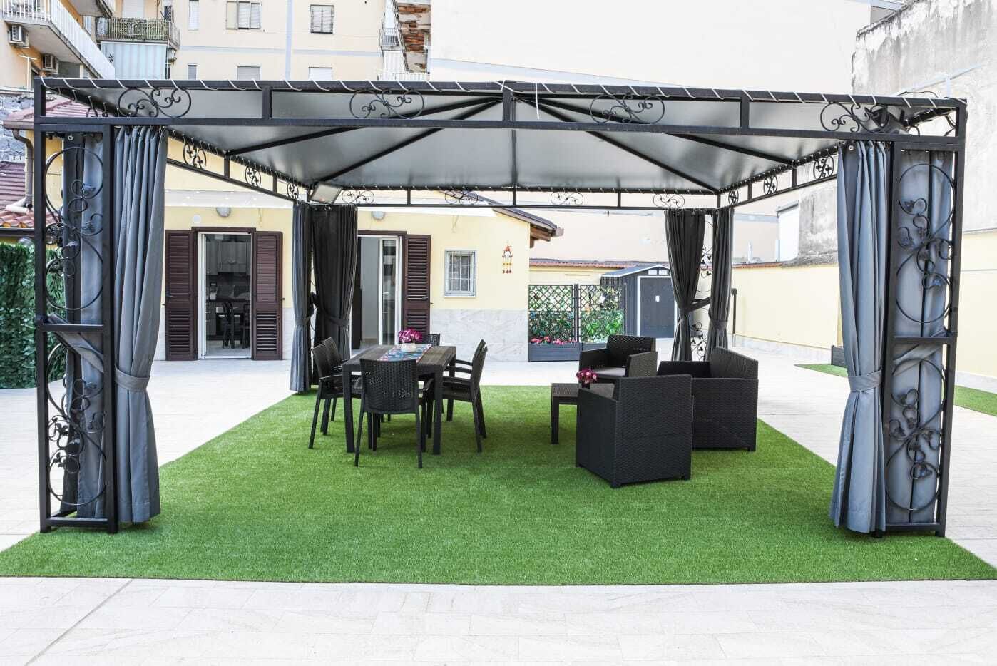 A covered outdoor seating area with a table and chairs, surrounded by a green lawn.