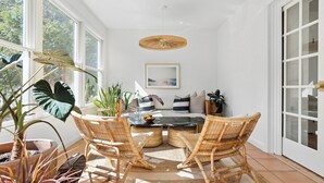 Una habitación iluminada por el sol con una mesa de comedor, sillas de mimbre, una lámpara colgante y un gran ventanal.