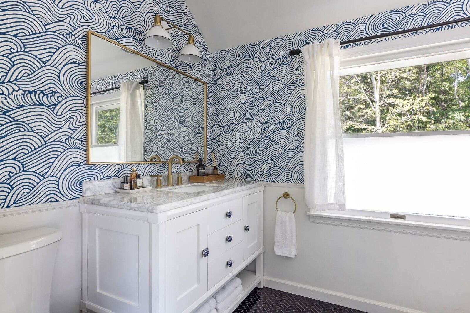 Une salle de bain avec un meuble-lavabo blanc, des robinetteries dorées, un grand miroir et une fenêtre donnant sur la verdure.
