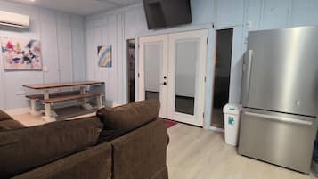 A living room with a sofa, a bench, a ceiling fan, a flat-screen TV, a refrigerator, and a trash bin.