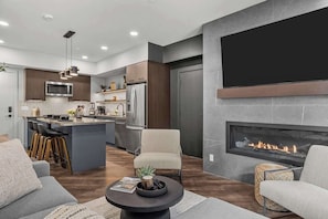 A modern kitchen with a central island, a dining area with bar stools, a fireplace, and a living room with sofas and a coffee table. - Spring Oasis: Brand New, Hot Tub/Pool, AC, Parking (Canmore)