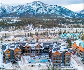 A multi-story hotel with a pool area, surrounded by snow-covered mountains and forested landscape. - Ascent Luxe Retreat | Brand-New 2BR (Canmore)