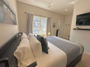 A hotel room with a bed, a desk, a chair, a window with curtains, a door, and a TV mounted on the wall. - Skyline Peaks Mountain Retreat - Studio (Canmore)