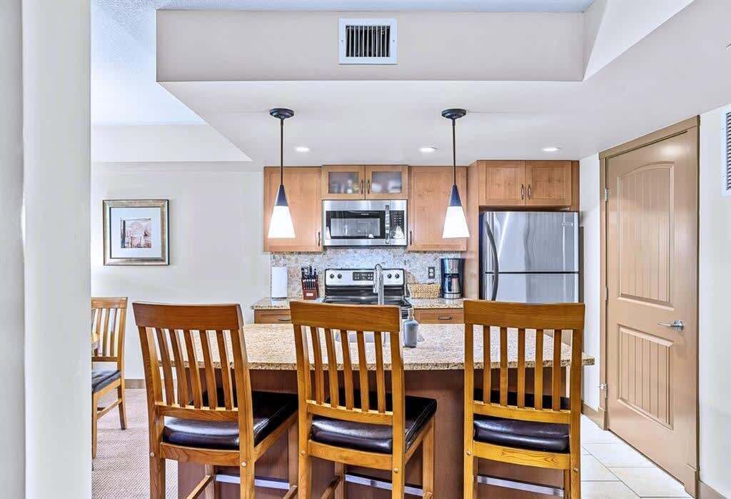 A modern kitchen with wooden cabinets, a stainless steel refrigerator, a microwave, and a gas stove. There are two pendant lights above the counter and a framed picture on the wall.