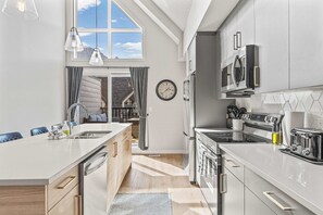 Ett modernt kök med en central köksö, rostfria vitvaror och utsikt över ett hus genom ett fönster.