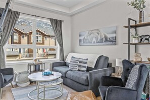 A living room with a grey sofa, a round coffee table, a view of a mountain house through a large window, and a bear picture on the wall. - Skyline Peaks Panorama Retreat (Canmore)