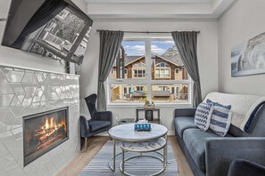 A living room with a fireplace, a sofa, a coffee table, and a view of a house through a window. - Skyline Peaks Panorama Retreat (Canmore)