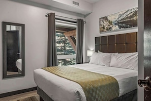 A hotel room with a bed, a large window with curtains, a headboard, a wall-mounted painting, and a mirror. - Rockies' Embrace- Pool, HotTub, Gym, BBQ, Parking (Canmore)
